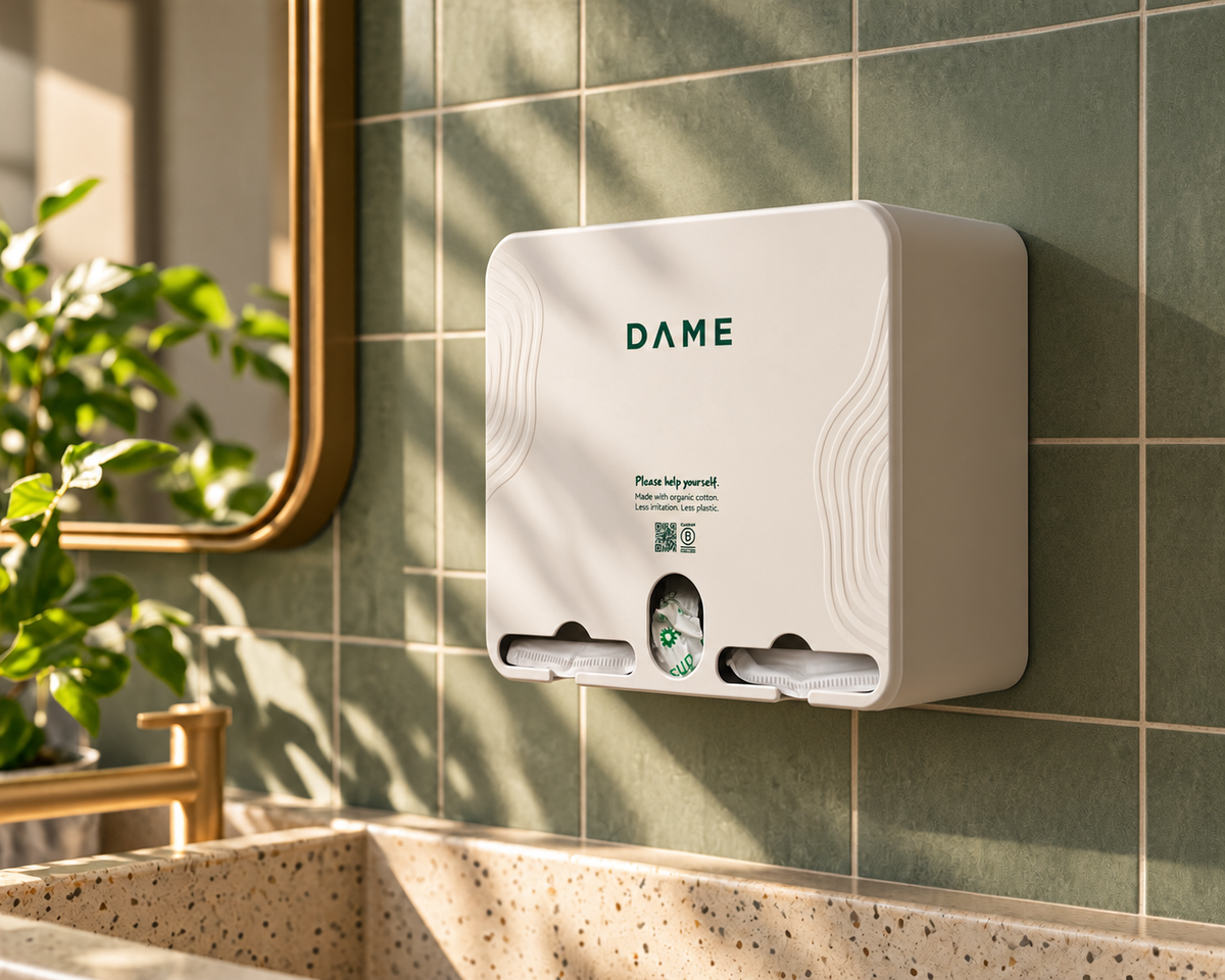 A wall-mounted DAME hand sanitizer dispenser in white, mounted on a green tiled bathroom wall above a speckled sink, with a plant and mirror visible.